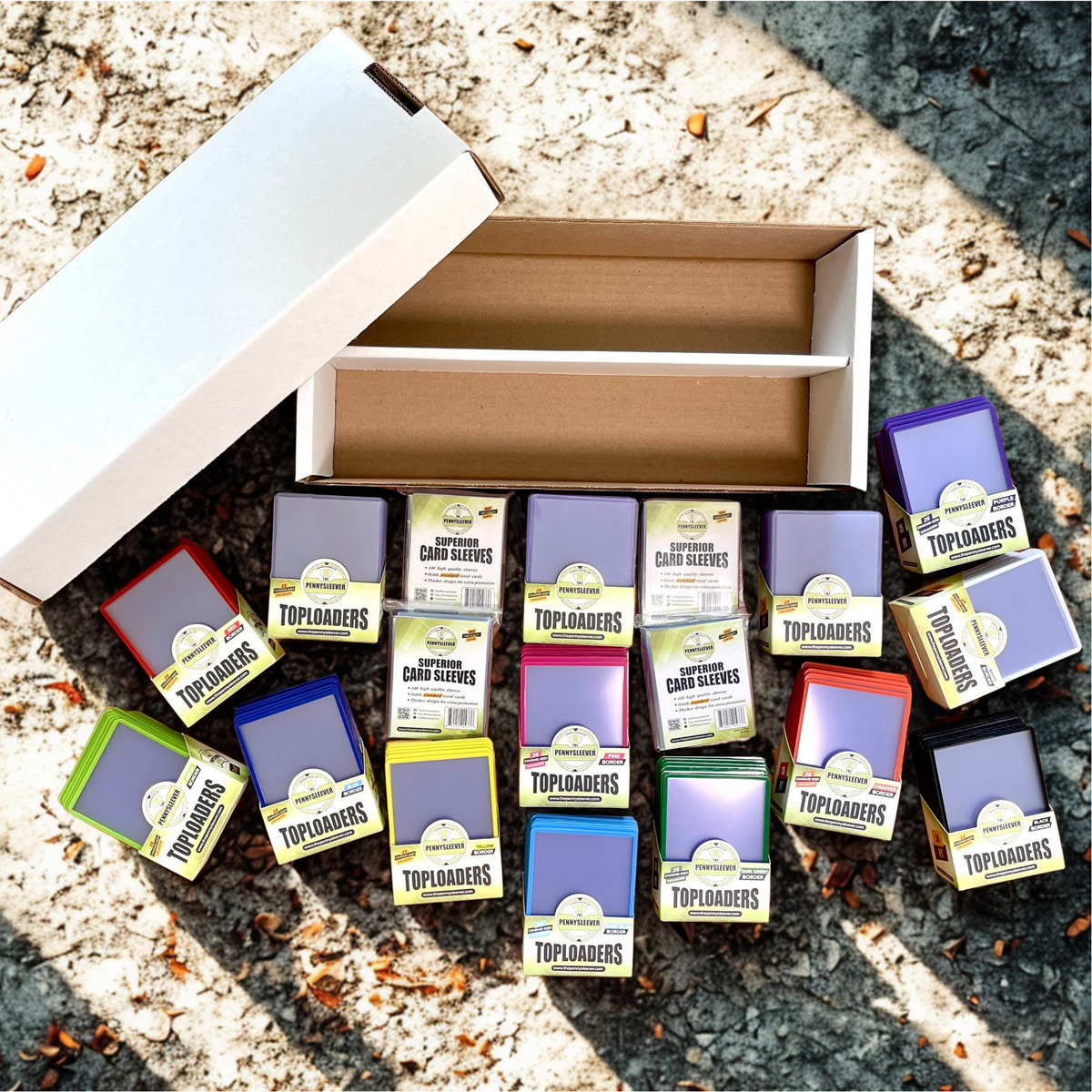 A display arrangement of 35pt Toploaders card sleeves, Superior Card Sleeves, and Color Frame 35pt Toploaders in multiple colors, including light green, pink, blue, yellow, black, purple, white, dark green, orange, light blue, and red displayed alongside an open two-row collector's cardboard box, arranged on an outdoor gravel surface with sunlight and shadows.