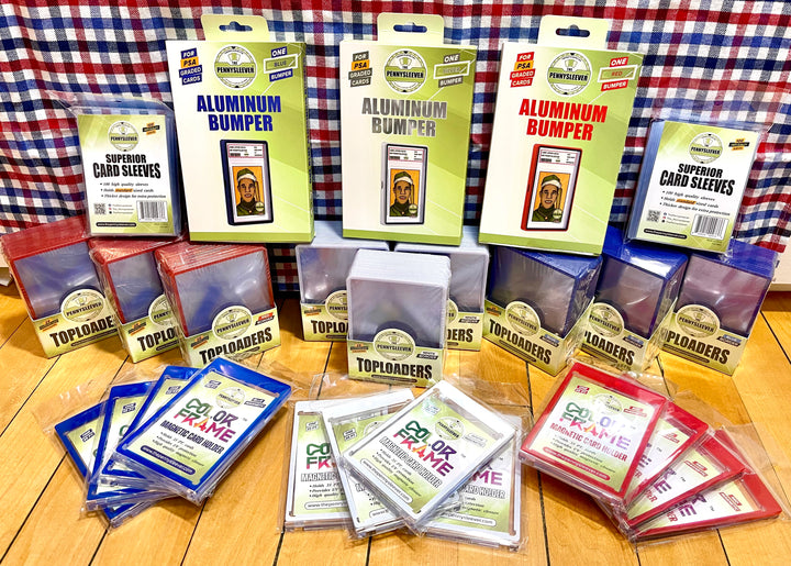 A collection of card protection products including aluminum bumpers, top loaders, and card sleeves in multiple colors, arranged on a wooden floor in front of a red, white, and blue plaid backdrop.